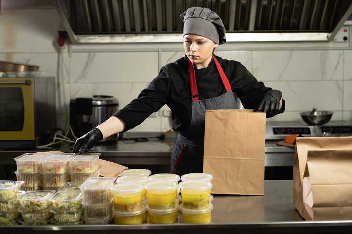 Female chef wearing an apron and gloves putting disposable plastic food containers into a disposable paper bag for delivery during a working day in a virtual kitchen that adheres to IWA 40:2022.