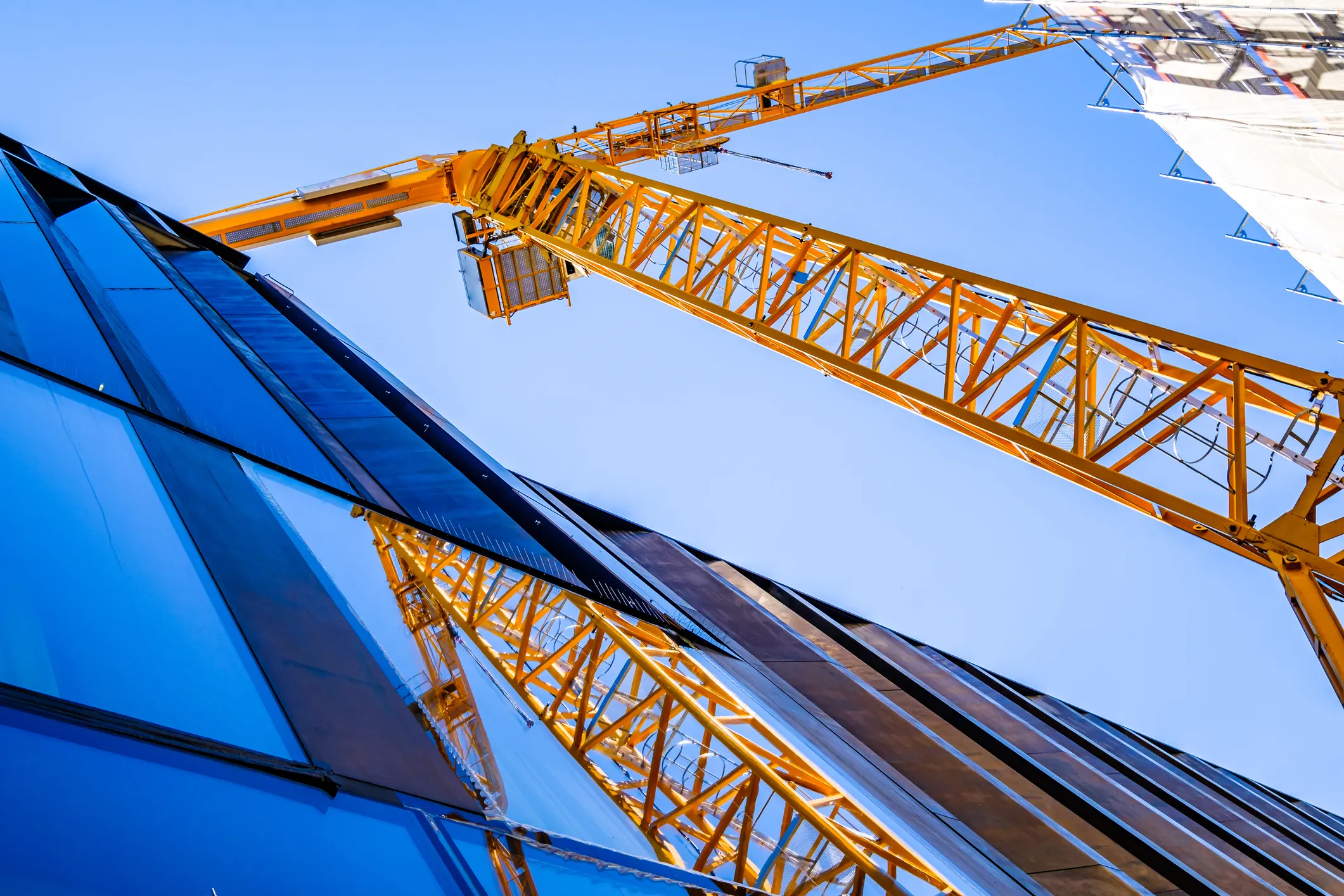 Building being constructed with the 2024 IBC and a bright yellow tower crane reflected in windows beside blue sky.