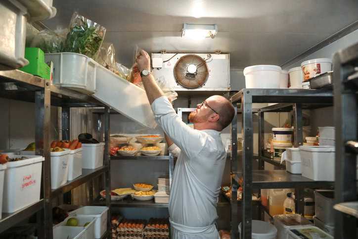 Bearded head chef at gourmet restaurant checking ingredients in walk-in refrigerator shelves.