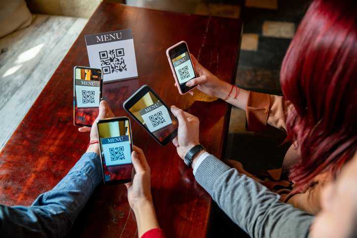 Group of people eating out at a restaurant who are scanning the menu with QR codes, adhering to ISO/IEC 18004:2024, on their cell phone cameras.