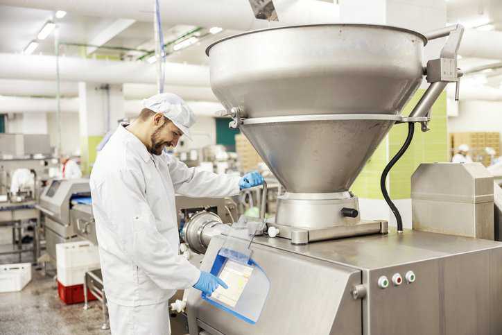 A meat industry laborer is managing meat grinding machine and pressing on dashboard in a commercial food factory with commercial food equipment adhering to NSF/ANSI 4-2024.
