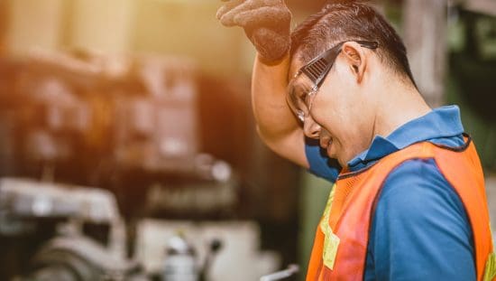 Worker in safety vest and glasses wiping the sweat off his brow from the machine heat.