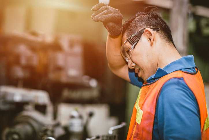 Worker in safety vest and glasses wiping the sweat off his brow from the machine heat.
