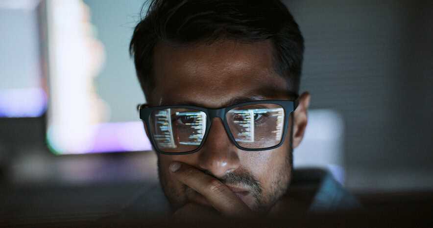 Coder staring at computer screen code, with eyeglasses reflecting screen in the dark.