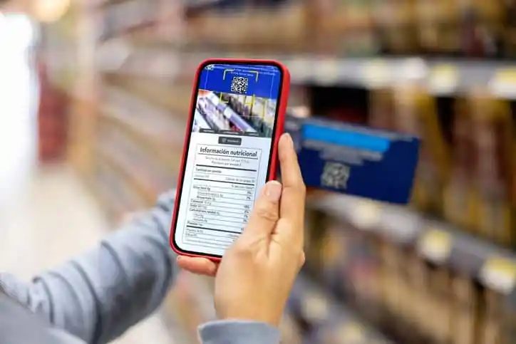 Close-up on a woman shopping at the supermarket and scanning a product barcode label, which adheres to ISO/IEC 15415:2024, with her cell phone to view the nutritional information.