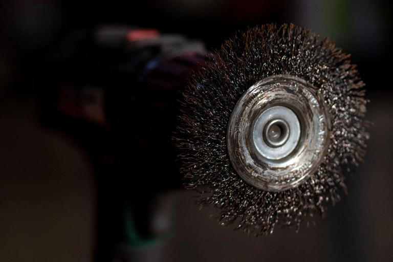 Steel power-driven brush, adhering to ANSI B165.1-2026, that is cleaning off old metal sheet with rust.