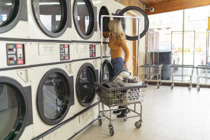 A young adult woman is pulling her clean laundry out of a washing machine at a laundromat, which has laundry equipment that adheres to ANSI Z8.1-2016.