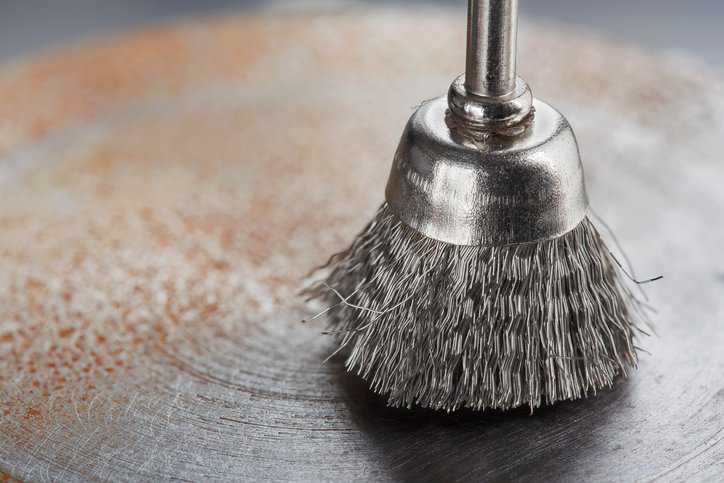 Steel power-driven brush, adhering to ANSI B165.1-2019, that is cleaning off old metal sheet with rust.