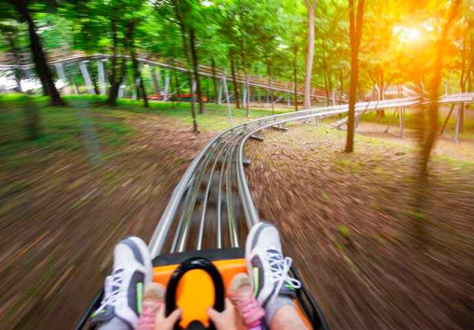 Father and daughter on a summer toboggan run, adhering to ISO 19202-1:2017 safety specifications, in a lush green forest.
