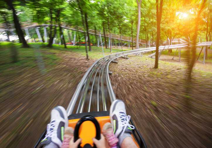 Father and daughter on a summer toboggan run, adhering to ISO 19202-1:2017 safety specifications, in a lush green forest.