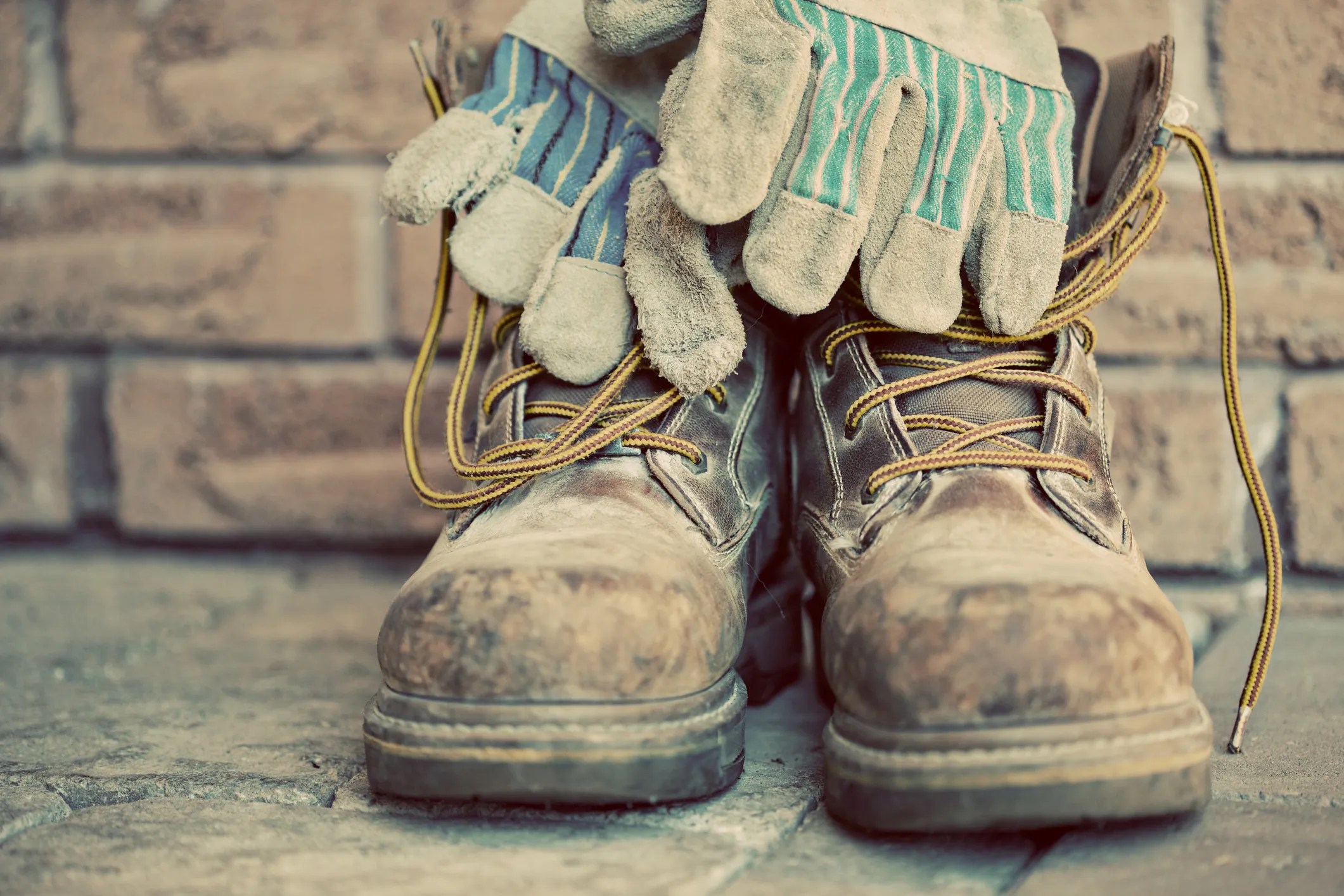 Old brown steel toe boots with work gloved in front of brick wall with sepia tone to show old ANSI Z41 standard.