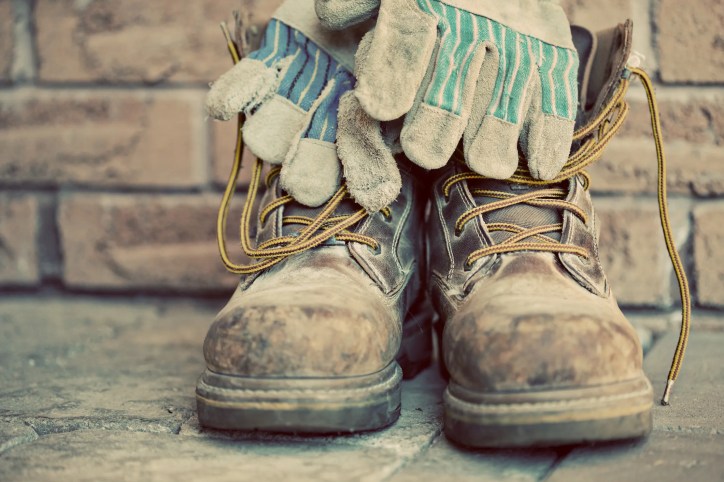 Old brown steel toe boots with work gloved in front of brick wall with sepia tone to show old ANSI Z41 standard.