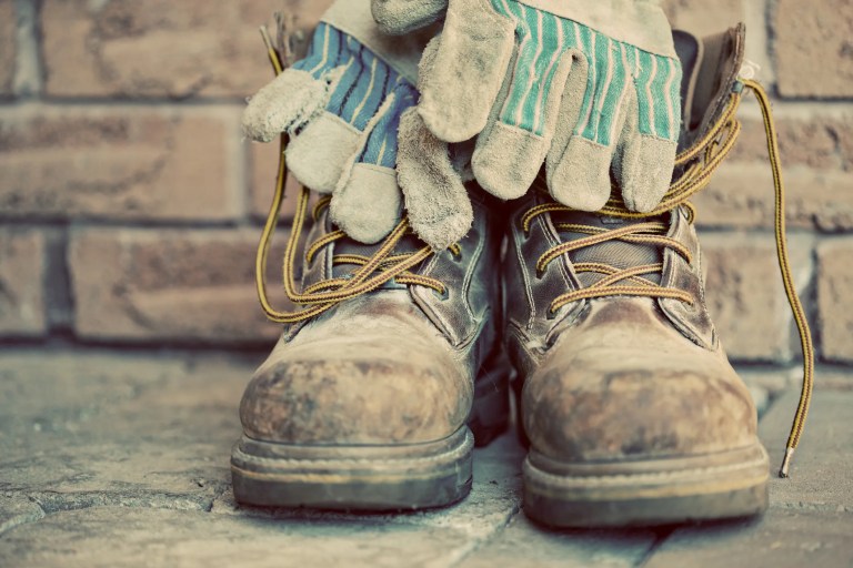 Old brown steel toe boots with work gloved in front of brick wall with sepia tone to show old ANSI Z41 standard.