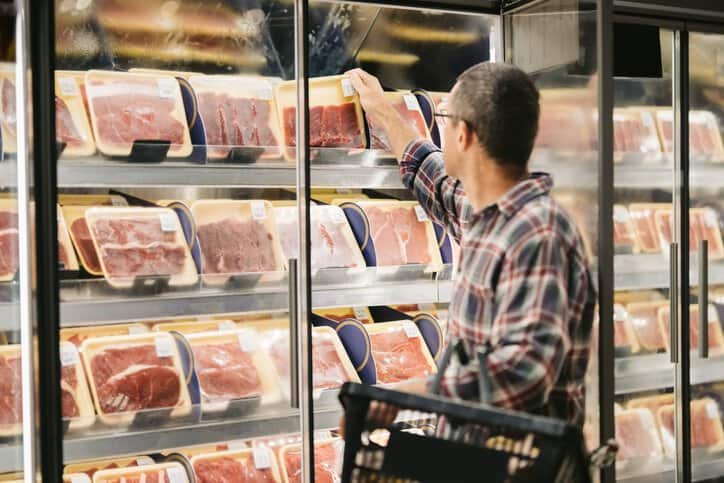 Male customer with a shopping basket taking meat from a fridge at local supermarket while examining its nutrition and quality claims, which adhere to ASTM F2925-11(2018).