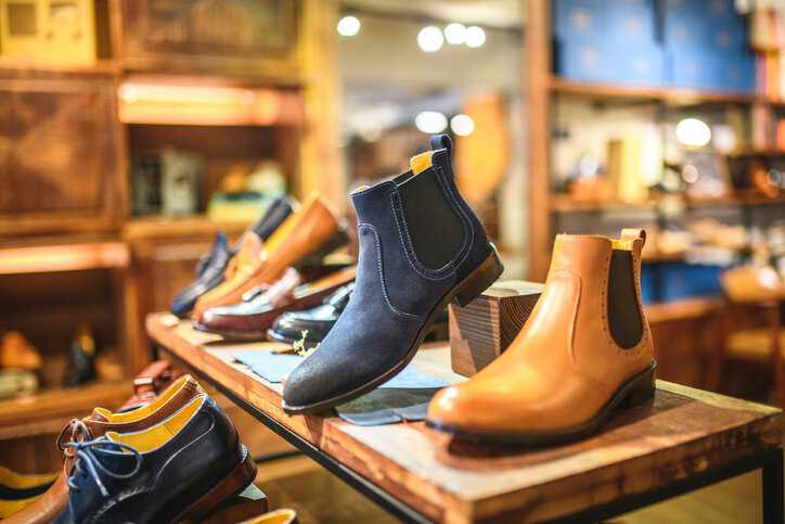 Close-up shot of blue and caramel color leather boots and shoes for men on wooden shelves that adhere to ISO 9407:2019 footwear sizing specifications.