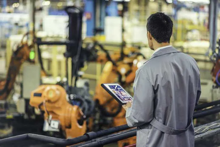 Rear view of an engineer in a gray coat examining quality control and compliance of autonomous robotic arms to ISO 10218-2:2025.