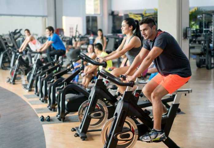 Happy group of people training at the gym cycling on stationary exercise bikes that comply with ISO 20957-5:2016.