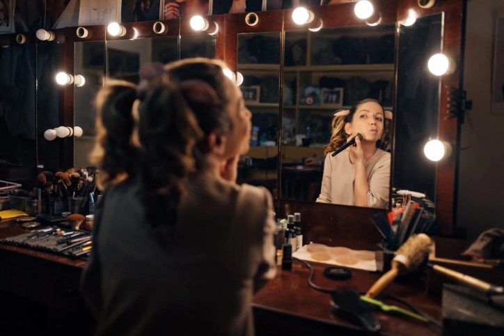 A Broadway dancer using a theatrical makeup mirror with lighting compliant to theatrical makeup mirror with lighting that adheres to ANSI E1.55 – 2016 (R2021).