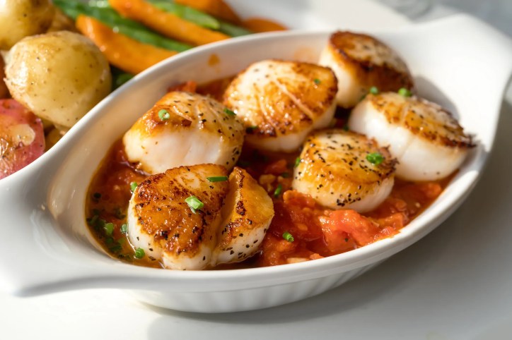 Beautifully seared golden-brown wet scallops prepared in tomato sauce, adhering to food safety standards.