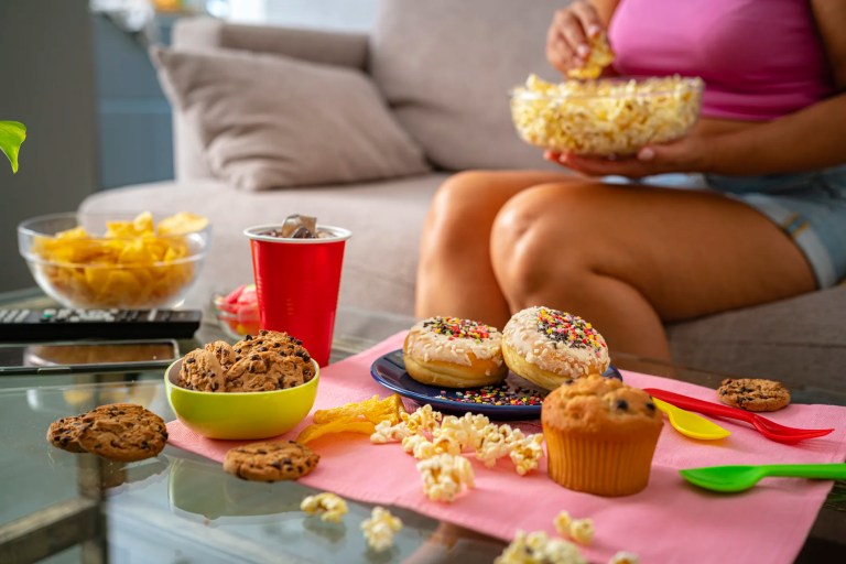 Woman on couch eating at risk of obesity and diabetes due to her unhealthy consumption of ultra-processed foods (UPFs), including soda, hamburger, and donuts.
