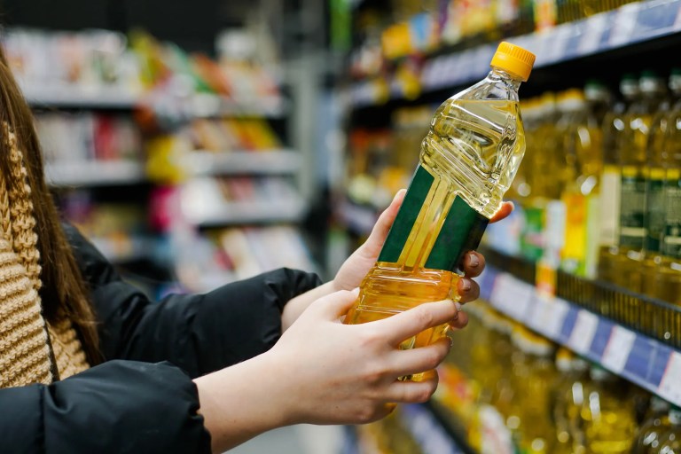 Woman in a grocery store examining various seed oils like sunflower, canola, and soybean oils in glass bottles, highlighting the variety of oils discussed in the seed oil debate.