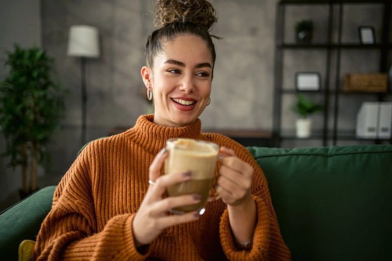 A young woman on her couch, who is drinking her morning cup of coffee experiencing the effects of drinking coffee on an empty stomach: acid production, cortisol stimulation, and blood sugar disruption.