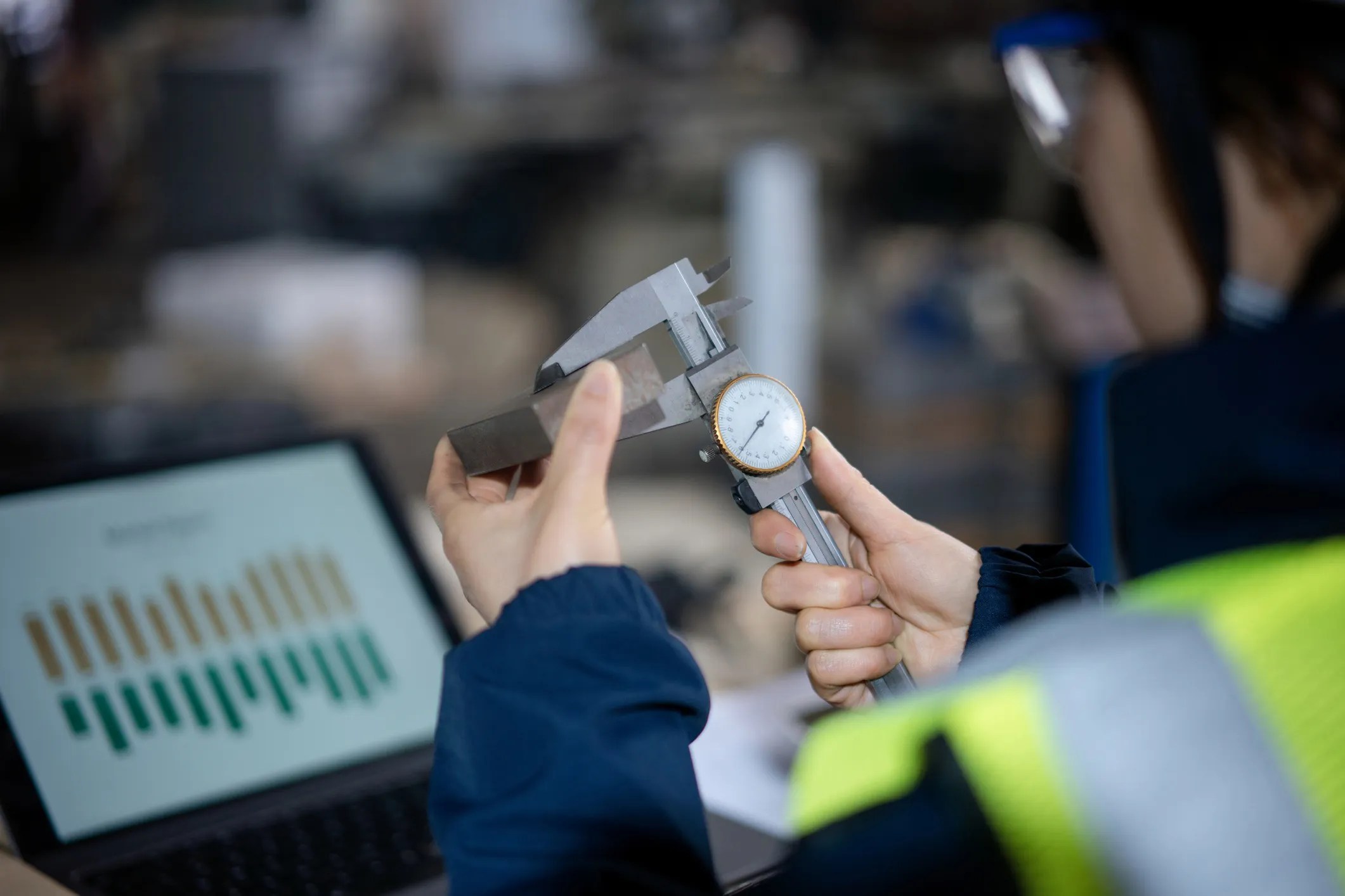 Female engineer using vernier calipers to measure metal parts in a manufacturing setting, assuring precise measurement and quality control, adhering to the ISO 10012:2026 standard for measurement management systems.