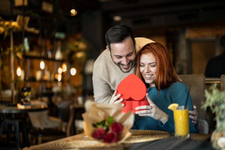 Man presenting his girlfriend with a Valentine’s Day gift and a bouquet of red roses, celebrating love and affection.