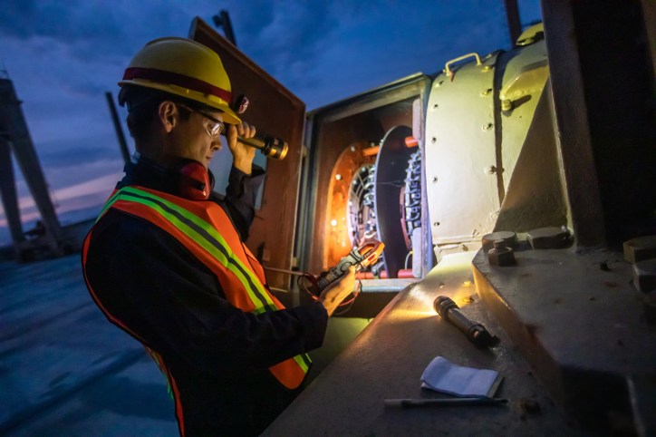 Outside under the darkness of night, a PPE protected electrician examines power plant wires with a flashlight.