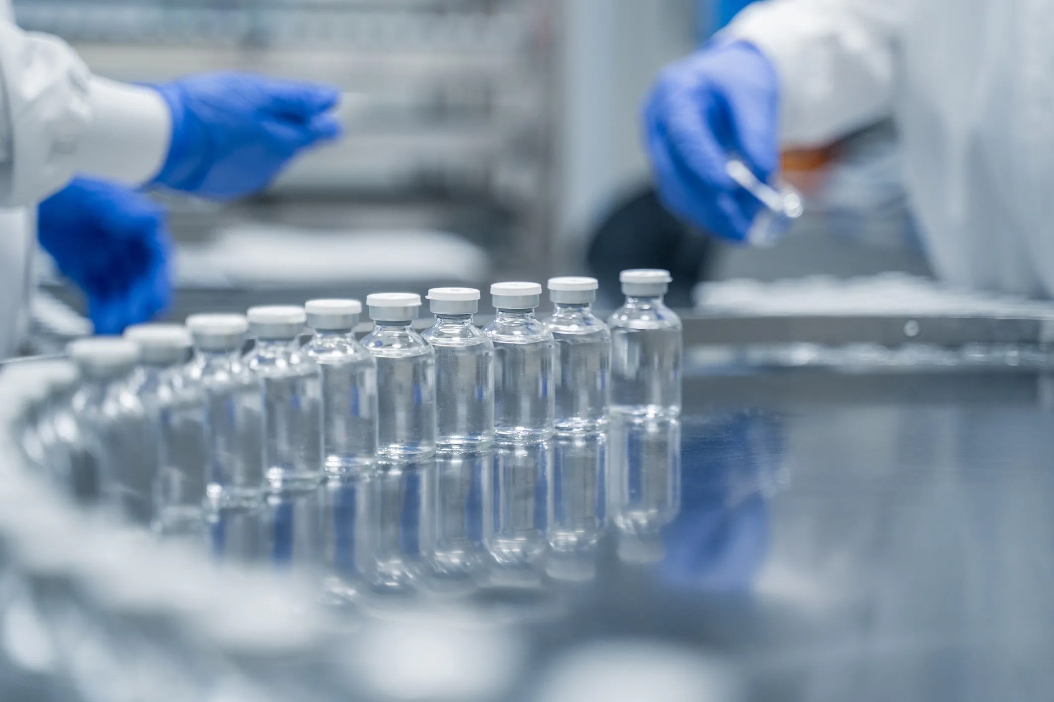 24 medicine vials moving along assembly line with clear liquid in front of gloved hands