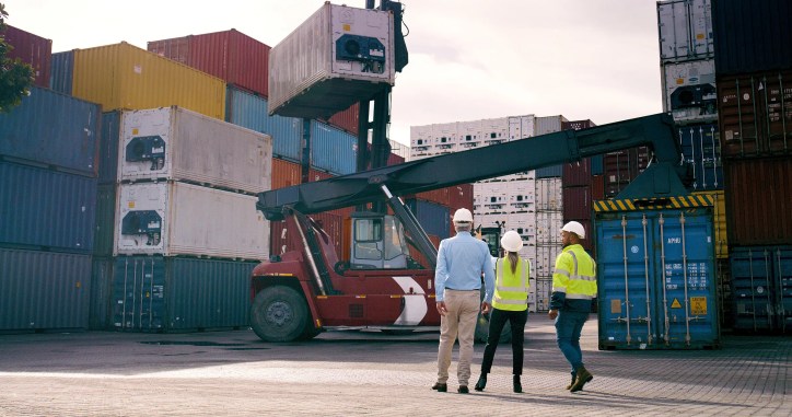 3 trusted accreditation professionals in hardhats and PPE watching colorful intermodal shipping containers moving.