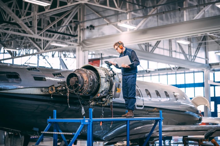 Engineer inspecting a commercial aircraft during manufacturing while technicians assemble aerospace components, illustrating AS9100 quality management and global aerospace supply chains.